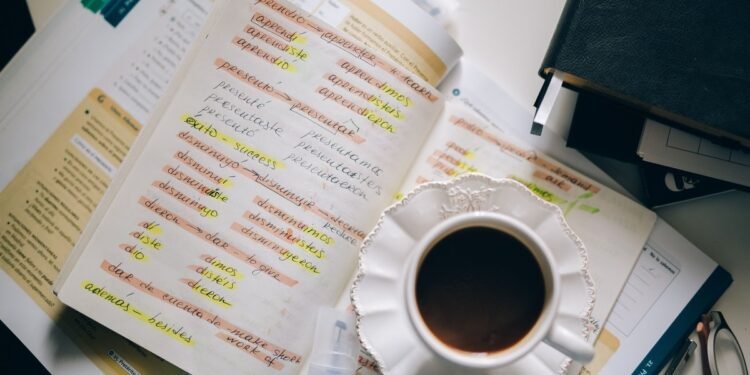 Caderno com anotações de conjugação verbal em espanhol, xícara de café ao lado, sobre uma mesa de estudo.