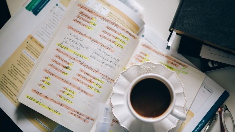 Caderno com anotações de conjugação verbal em espanhol, xícara de café ao lado, sobre uma mesa de estudo.