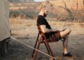 Homem sentado numa cadeira dobrável ao lado de uma barraca de acampamento, trabalhando num notebook, com montanhas de deserto ao fundo.
