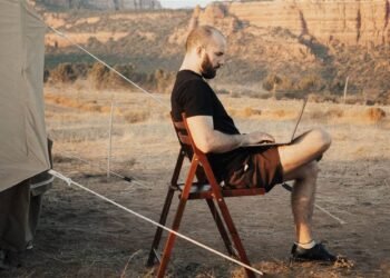 Homem sentado numa cadeira dobrável ao lado de uma barraca de acampamento, trabalhando num notebook, com montanhas de deserto ao fundo.