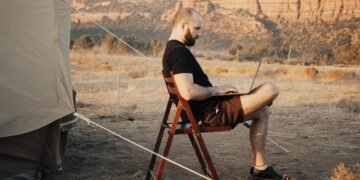 Homem sentado numa cadeira dobrável ao lado de uma barraca de acampamento, trabalhando num notebook, com montanhas de deserto ao fundo.