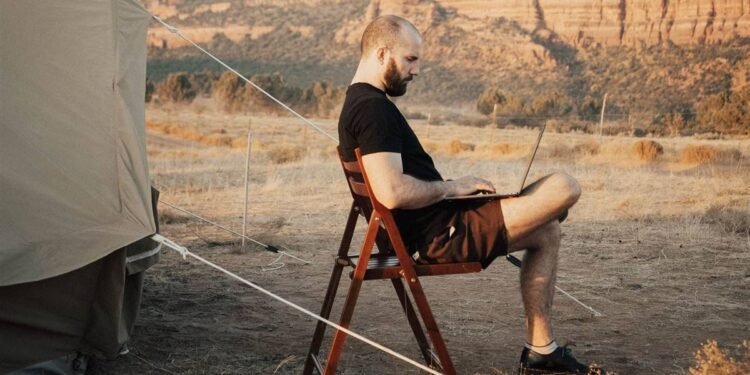 Homem sentado numa cadeira dobrável ao lado de uma barraca de acampamento, trabalhando num notebook, com montanhas de deserto ao fundo.