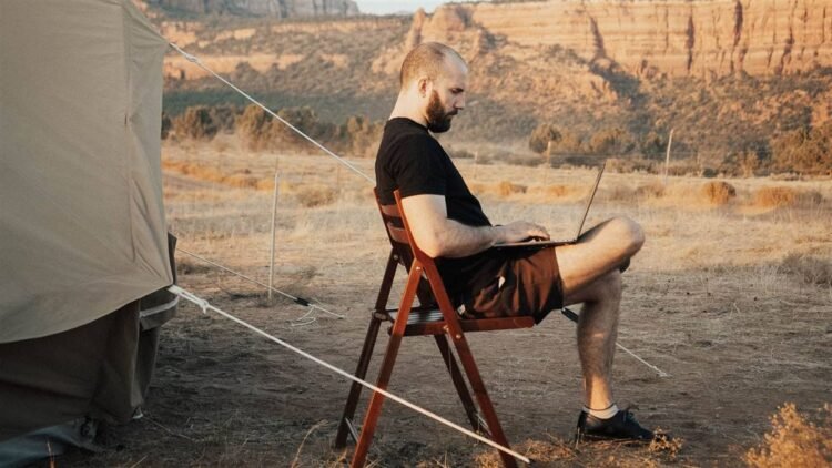 Homem sentado numa cadeira dobrável ao lado de uma barraca de acampamento, trabalhando num notebook, com montanhas de deserto ao fundo.