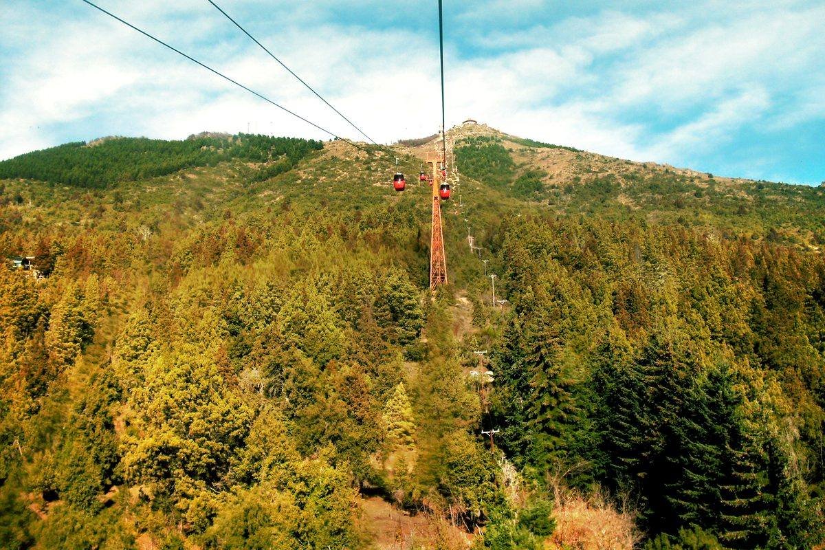 Teleférico do Cerro Otto subindo entre a mata, com bondinhos vermelhos e vista da montanha.