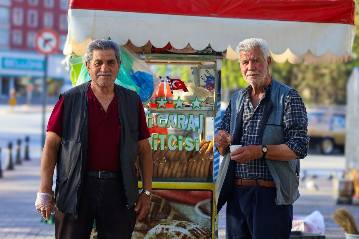 Dois vendedores de rua sorrindo ao lado de um carrinho de simit, em uma praça.