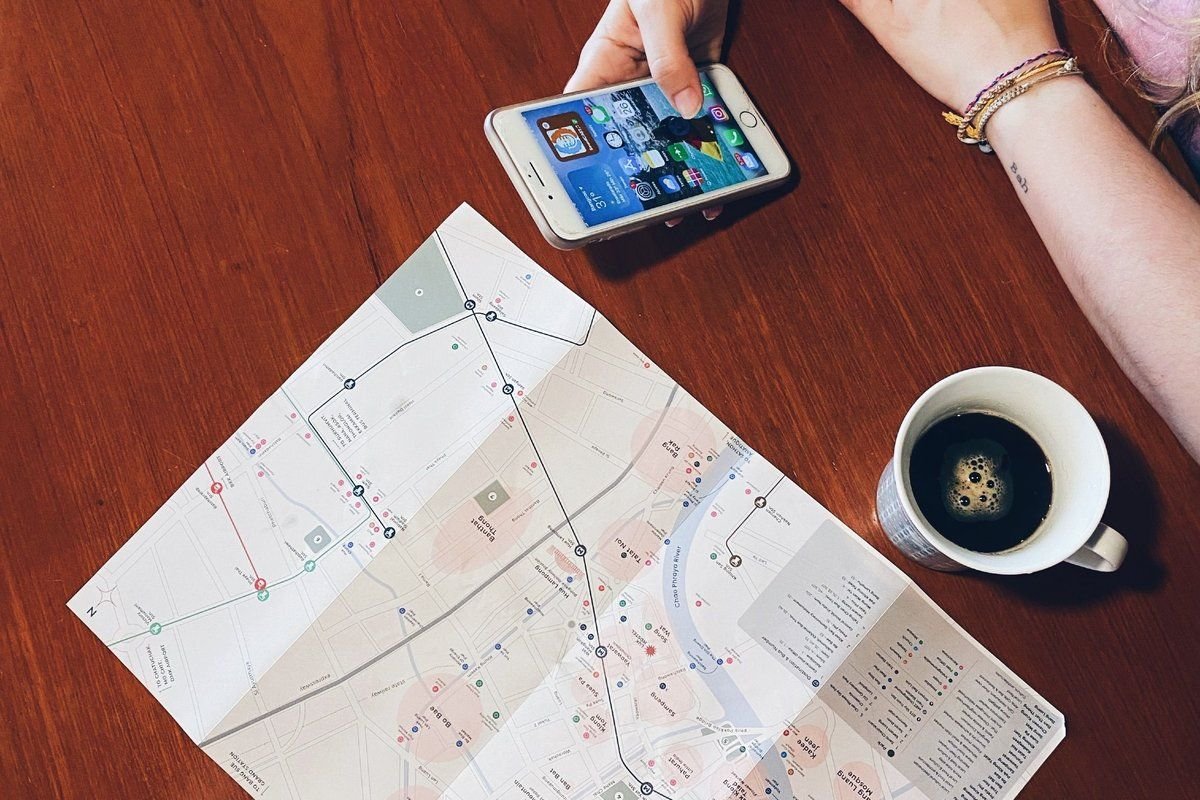 Mão segurando um celular com aplicativo aberto, sobre um mapa impresso, com uma xícara de café ao lado.