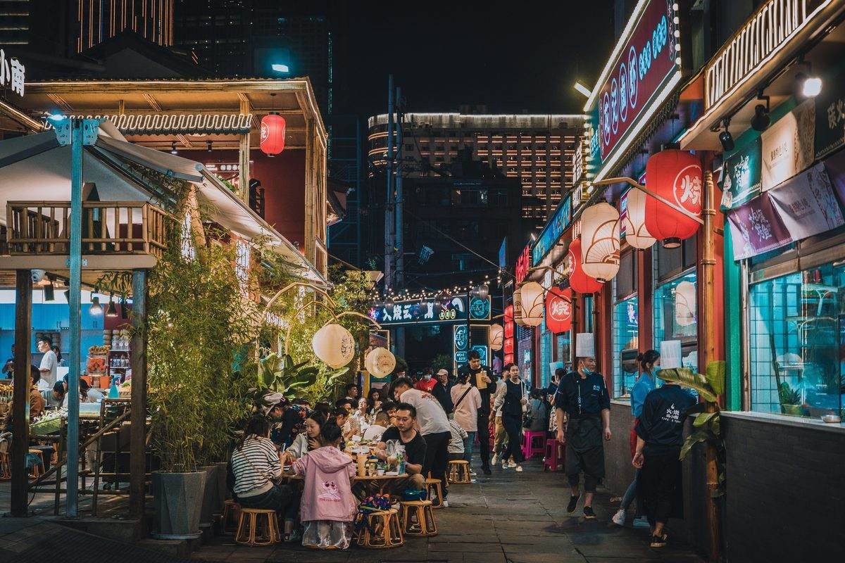 Mercado de comida de rua iluminado à noite, com pessoas sentadas em mesas comendo.