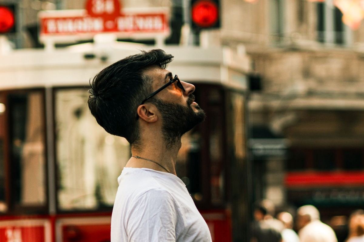 Homem de óculos escuros observa o ambiente ao redor, com bandeirinhas coloridas e um bonde histórico ao fundo.
