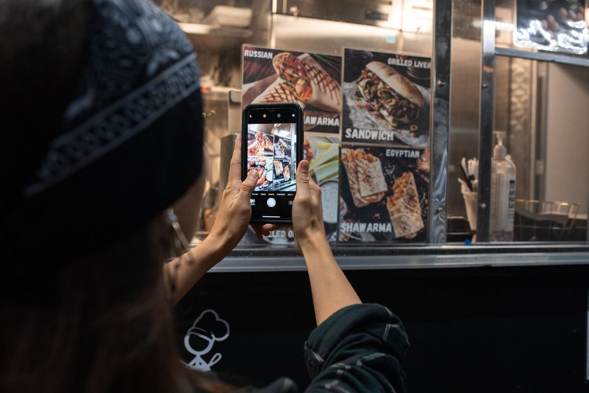 Pessoa fotografando o cardápio de uma barraca de comida de rua com o celular, à noite.