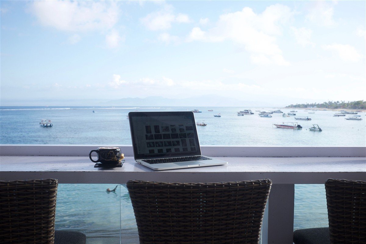 Notebook e xícara de café sobre uma mesa, com vista do mar ao fundo.