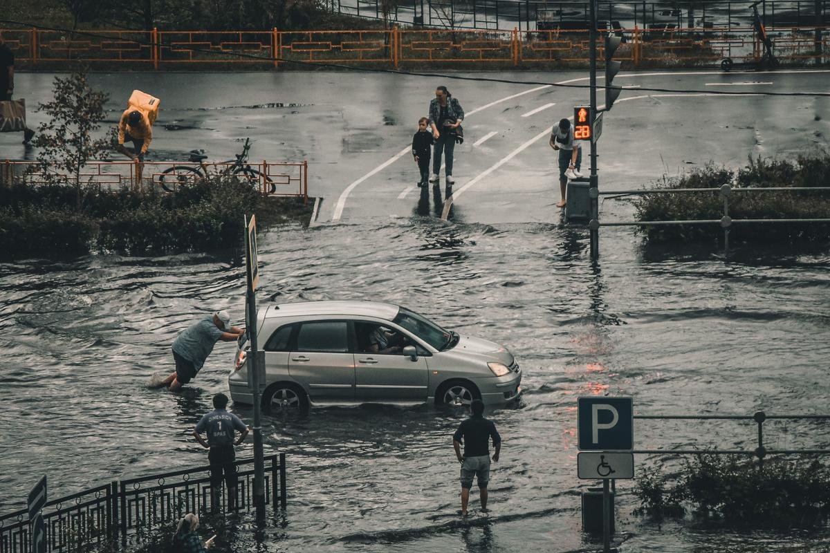 Rua alagada, com carro parado na água e pedestres passando pela enchente.
