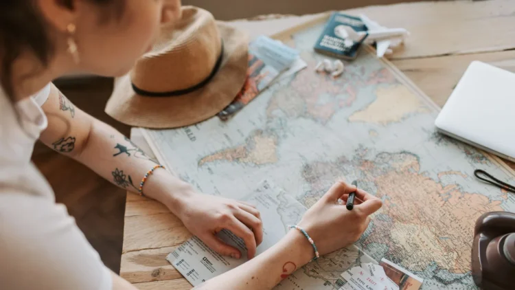 Pessoa inclinada sobre um mapa-múndi aberto numa mesa de madeira, segurando uma caneta sobre o mapa com uma das mãos e um folheto com a outra; ao redor, um chapéu de palha, um passaporte com um avião de brinquedo, um notebook fechado e uma câmera fotográfica.