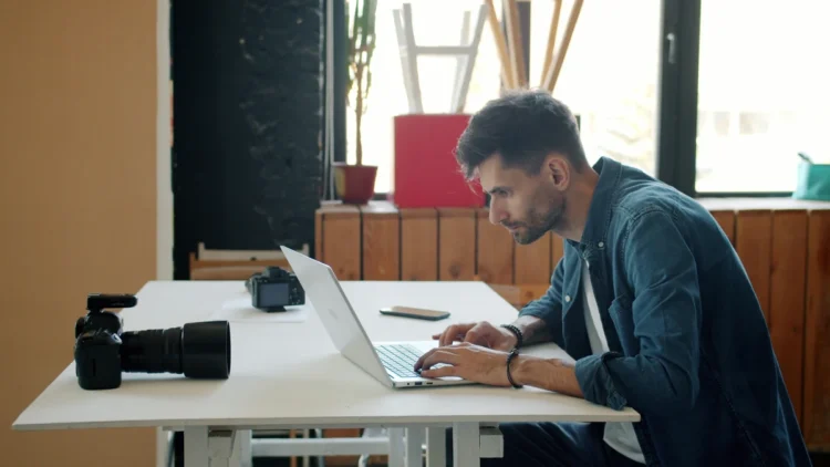 Fotógrafo edita imagens em um laptop, com uma câmera sobre a mesa.