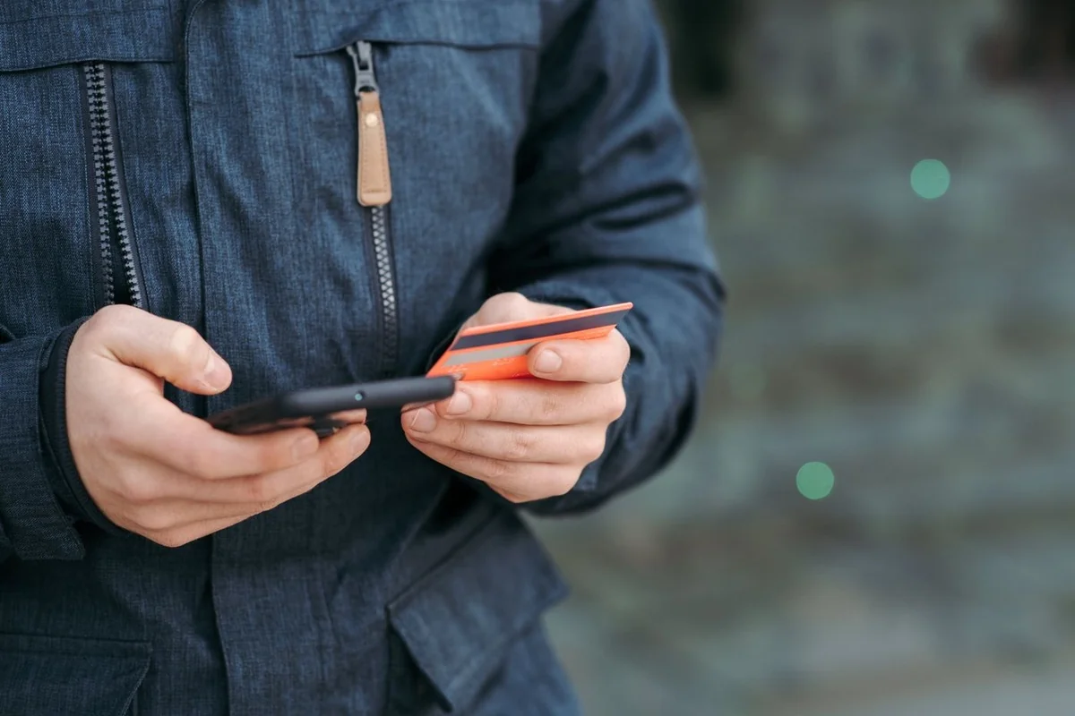Pessoa de jaqueta escura segura dois cartões de crédito em uma das mãos e um celular na outra, conferindo a tela na rua.