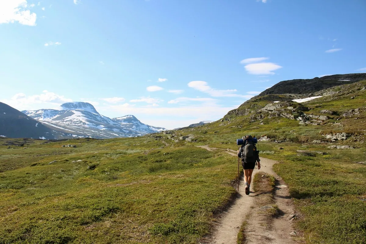 Caminhante com mochila cargueira e bastão de trekking sobe trilha de terra em campo aberto de altitude, com montanha coberta de neve ao fundo.