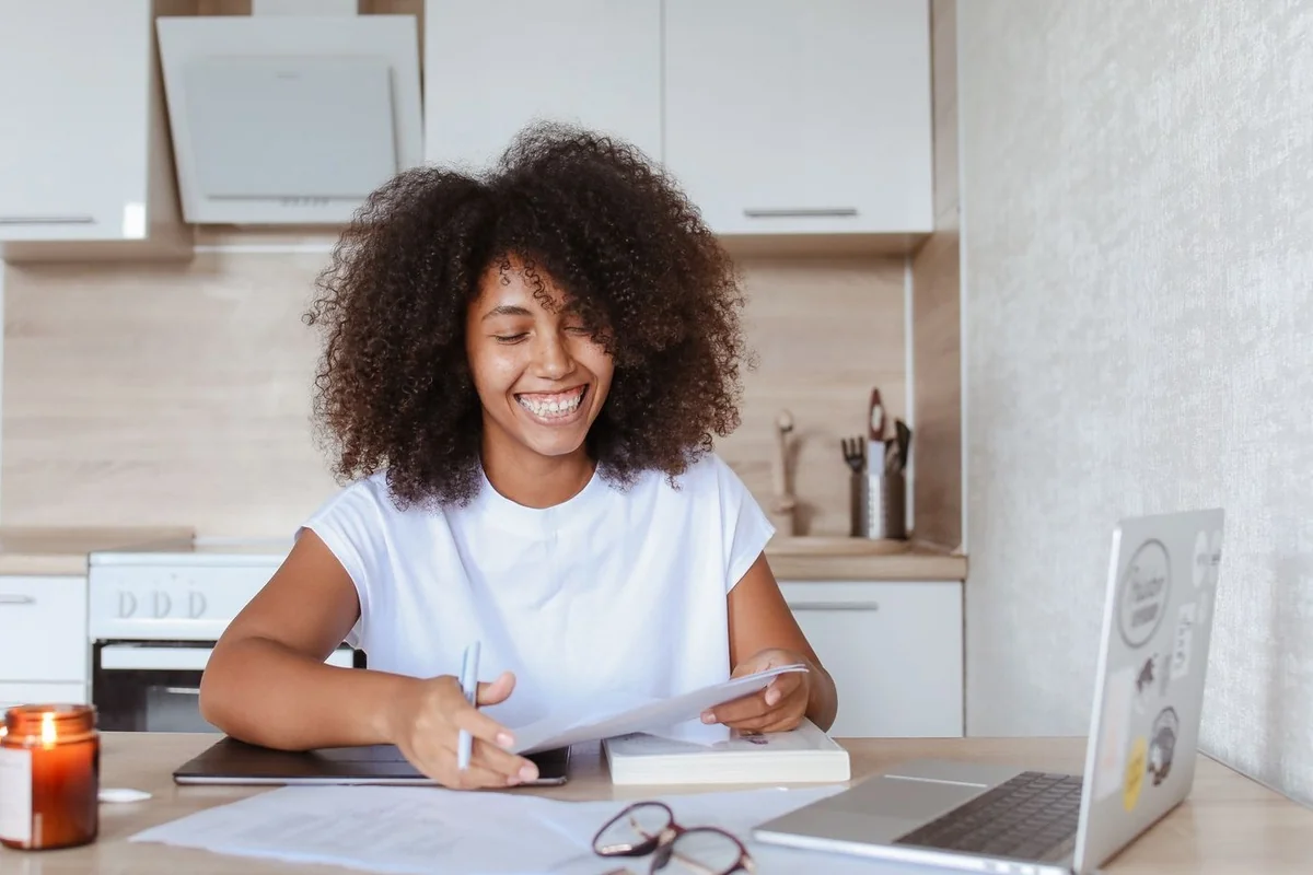 Mulher sorridente, sentada a uma mesa de cozinha, segura uma folha impressa com as duas mãos e uma caneta na direita, com papéis, óculos, um caderno e um notebook aberto sobre a mesa.